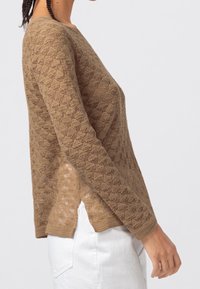 Beżowy sweter z dzianiny o fakturowanym wzorze, z okrągłym dekoltem i długimi rękawami. Dodatkowe detale to boczne rozcięcia, zestawione z jasnymi spodniami.