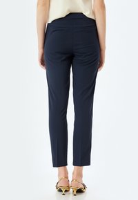 Pantaloni aderenti blu navy in tessuto liscio, con due tasche posteriori e un design sartoriale, abbinati a scarpe con tacco dorato.