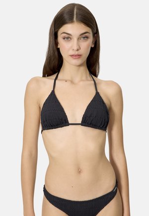 Mujer con cabello largo y castaño, vistiendo un bikini negro texturizado, de frente contra un fondo liso y claro.