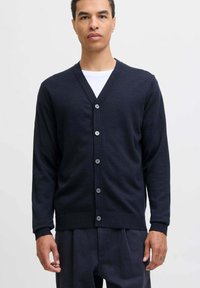 Uomo che indossa un cardigan blu navale con bottoni sopra una camicia bianca, in piedi di fronte a uno sfondo chiaro e semplice, rivolto in avanti.