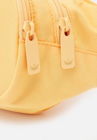 Žlutý textilní sáček s texturovaným povrchem, vybavený dvojitými zipy a zipovými táhly s logem, vše v jednotné barevné paletě.