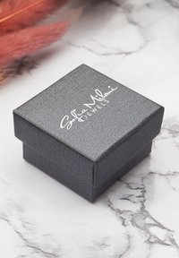 Svart texturerad smyckeslåda med kvadratisk form och en vit logotyp på locket. Ligger på en ljus marmor yta.