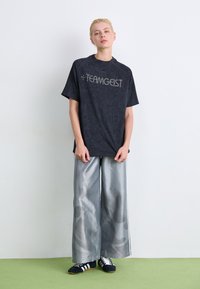 Zwarte T-shirt met korte mouwen met een textuurpatroon van bladeren en zilveren "TEAMGEIST" letters, gecombineerd met brede, glanzende grijze broek en zwarte sneakers.