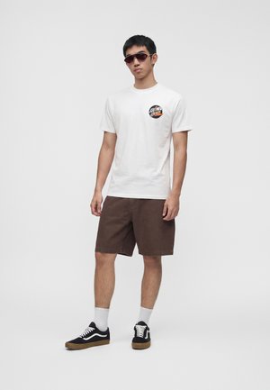 Άνδρας που φοράει λευκό γραφικό t-shirt, καφέ σορτς μήκους μέχρι το γόνατο, μαύρα αθλητικά με λευκά κορδόνια, λευκές κάλτσες και σκούρα γυαλιά ηλίου, στέκεται μπροστά σε λευκό φόντο.