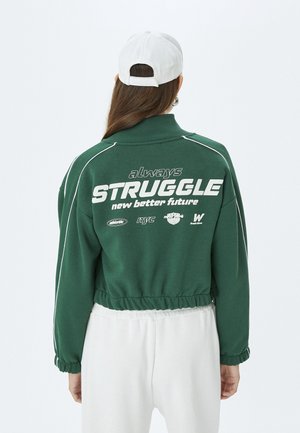 Sweatshirt court vert avec des accents blancs et un ourlet élastiqué. Un grand texte blanc au dos indique "toujours LUTTER pour un avenir meilleur."