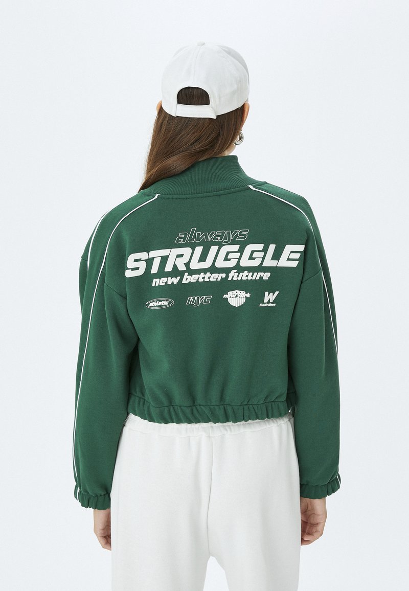 Sudadera verde recortada con detalles blancos y dobladillo elástico. Gran texto blanco en la parte posterior que dice "siempre LUCHA por un futuro mejor".