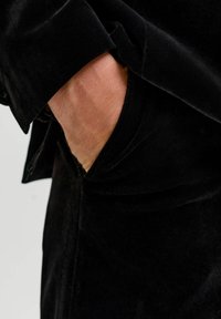 Čierny zamatový odev s prehnutím na manžete. Ruka je čiastočne viditeľná v vrecku, čo ukazuje hladkú textúru a elegantný dizajn.