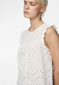 Blouse blanche sans manches avec broderie œillet, comportant un col rond et des épaules volantées, mettant en valeur un matériau léger et texturé.