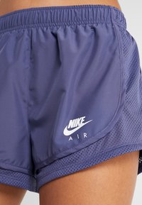 Lähikuva purppuraisista Nike Air -juoksushortseista, joissa on verkkomateriaali helman alueella, päällä lonkalla ja yläreidellä kirkasta taustaa vasten.