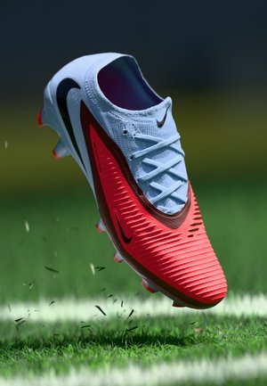 Nike nogometna kopačka s svijetloplavom gornjom površinom, crvenim teksturiranim dizajnom, crnim detaljima i narančastim udarcima, lebdi iznad trave s učinkom zamućenja pokreta.