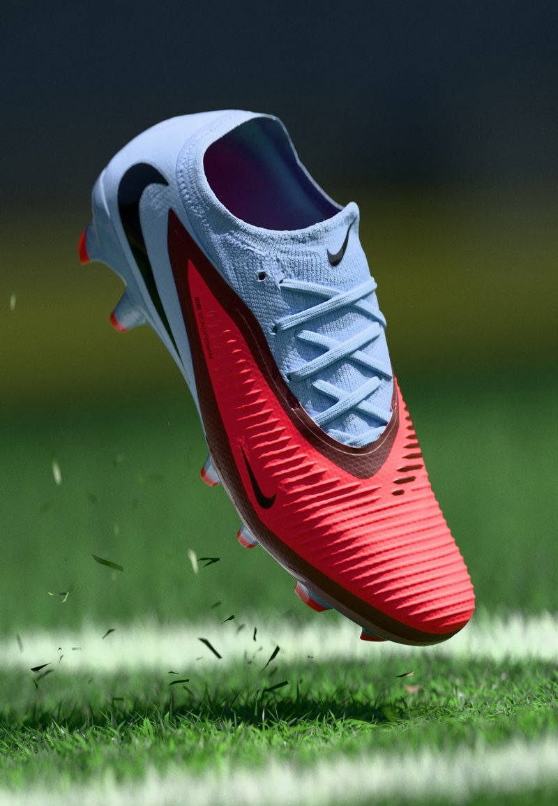 Botines de fútbol de Nike con un upper azul claro, diseño texturizado rojo, acentos negros y tacos naranjas, flotando sobre el césped con desenfoque de movimiento.