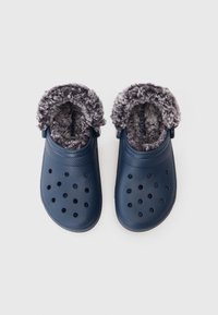 Blå slip-on træsko med sorte ventilationshuller og tykk grå pelsforing, designet til varme og komfort.