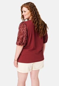 Chemise courte à manches courtes bordeaux avec un motif floral brodé texturé sur des manches bouffantes, associée à un short couleur crème. Tissu en coton lisse.