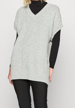 Femme portant un pull sans manches gris clair en maille torsadée avec col en V, par-dessus un haut à manches longues noir et un pantalon noir, debout devant un fond blanc.