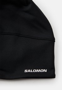 Svart pandebånd laget av elastisk stoff, har en glatt tekstur og sydd detaljer, med "SALOMON"-logoen i hvitt på fronten.