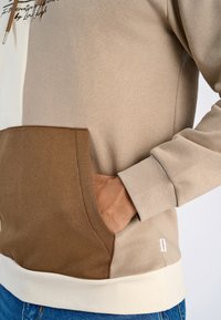 Beige hoodie met een color-block ontwerp met lichtbruine en donkerbruine accenten. Inclusief een kangoeroe pocket en ribgebreide manchetten.