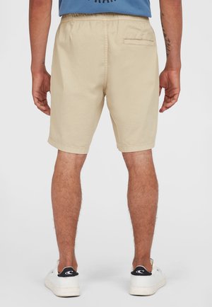 Homme portant un short beige et des baskets blanches, vu de dos, se tenant contre un fond clair uni.