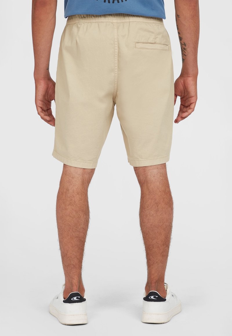 Man wearing beige shorts and white sneakers, shown from the back, standing against a plain light background.