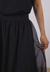 Rochie neagră cu un top ajustat și o fustă neagră cu straturi, din material transparent. Țesătura este netedă pentru partea de sus și curgătoare pentru fustă. Mână odihnindu-se pe fustă.