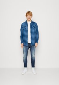 Camisa de cuadros azul con dos bolsillos en el pecho, sobre una camiseta blanca, combinada con jeans de mezclilla ajustados y desgastados y zapatillas blancas.