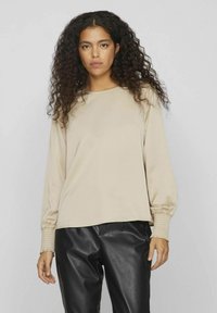 Beige långärmad topp med rund halsringning och elastiska ärmmuddar, i kombination med svarta faux läderbyxor. Slätt, lätt material.