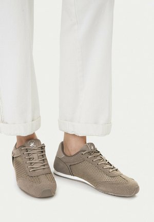 Piedi che indossano sneakers in mesh e pelle scamosciata taupe abbinate a pantaloni bianchi con risvolto su uno sfondo semplice.