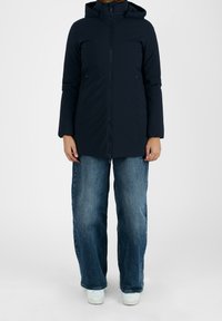 Personne portant une veste à capuche noire, un jean bleu à jambes larges et des baskets blanches, se tenant contre un fond blanc uni.