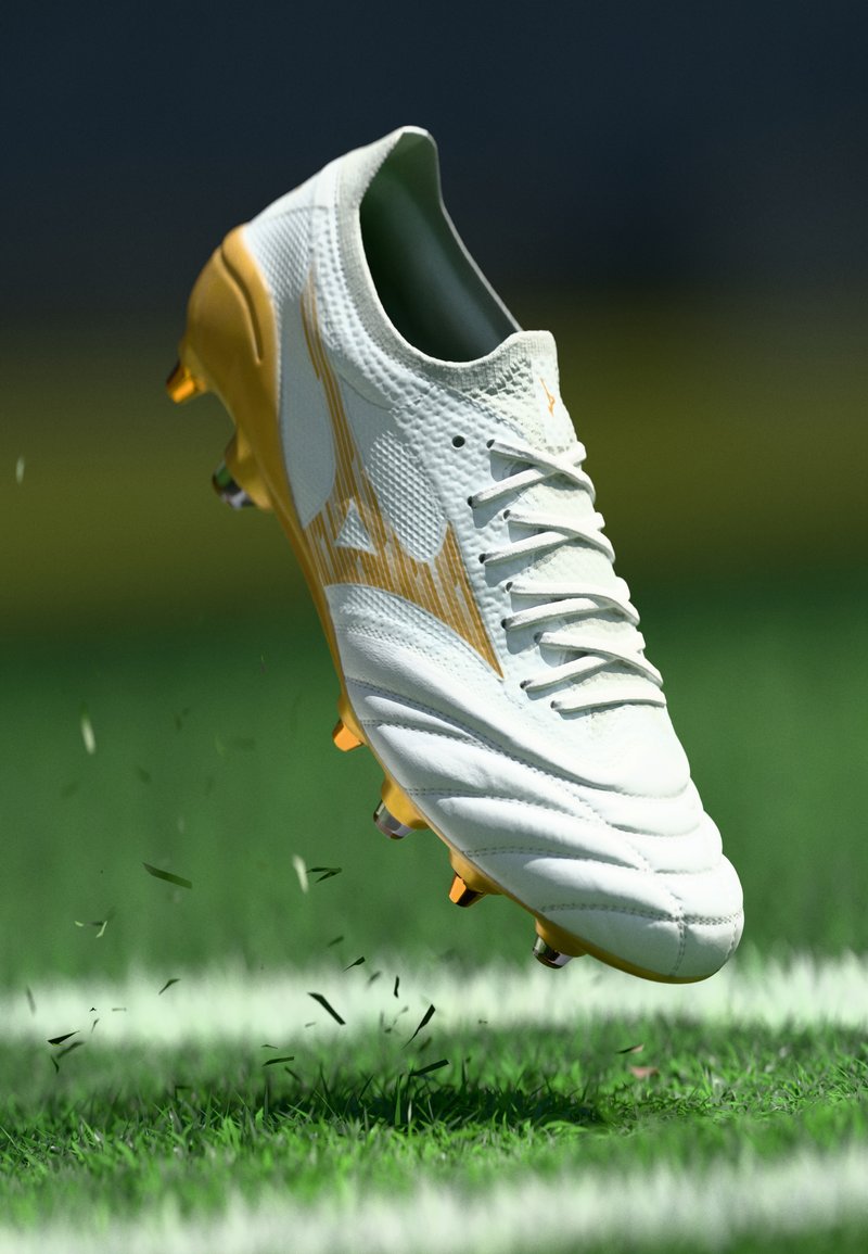 White soccer cleat with yellow accents, textured upper, and six molded studs, positioned above green grass, kicking up small debris.