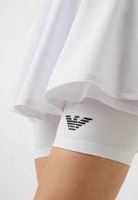 Balta plisētā svārku virsma ar baltām kompresijas šortiem, kuru rotā melns logotips ar spārniem, izgatavoti no viegla materiāla ērtībai un elastībai.