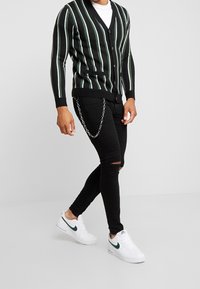 Cardigan a rayas negro y verde con botones, combinado con jeans negros desgastados que presentan un acento de cadena, y zapatillas blancas con logo verde.
