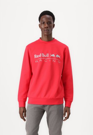 Mand iført rød crewneck-sweatshirt med "Red Bull Racing"-logo, kombineret med lysegrå jeans, stående foran en ensfarvet hvid baggrund.