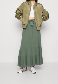 Veste bomber en satin vert olive portée sur un crop top blanc, associée à une jupe maxi verte à volants et des baskets beiges. Comprend des détails avec cordon de serrage.