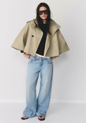 Kobieta ubrana w oversize'ową beżową, krótką pelerynę, czarny top, szerokie jasnoniebieskie jeansy, czarne sandały i duże ciemne okulary przeciwsłoneczne, stojąca z rękami w kieszeniach.