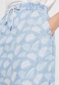 Světle modré šortky s bílým tropickým listovým vzorem, elastický pas s bílou tkaničkou a ruka osoby v kapse.