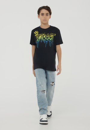 T-shirt nera con testo in stile graffiti "STREET" in giallo e blu, abbinata a jeans chiari strappati e sneaker bianche.