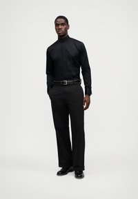 Camisa preta com colarinho alto, combinada com calças pretas e sapatos pretos polidos. Tecido suave com corte ajustado.