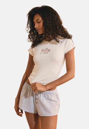 Femme aux cheveux bouclés portant un t-shirt blanc à manches courtes avec l’inscription "The Book Club" et un short à carreaux clairs avec des boutons.