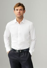 Chemise blanche à manches longues avec un col évasé, associée à un pantalon sombre et une ceinture noire avec des accents en argent. Finition en tissu lisse.