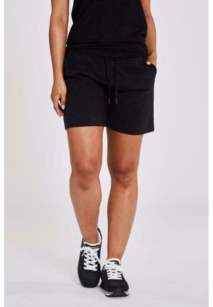 Personne portant un short noir à cordon, un haut noir et des baskets noires à lacets blancs, debout devant un fond uni.