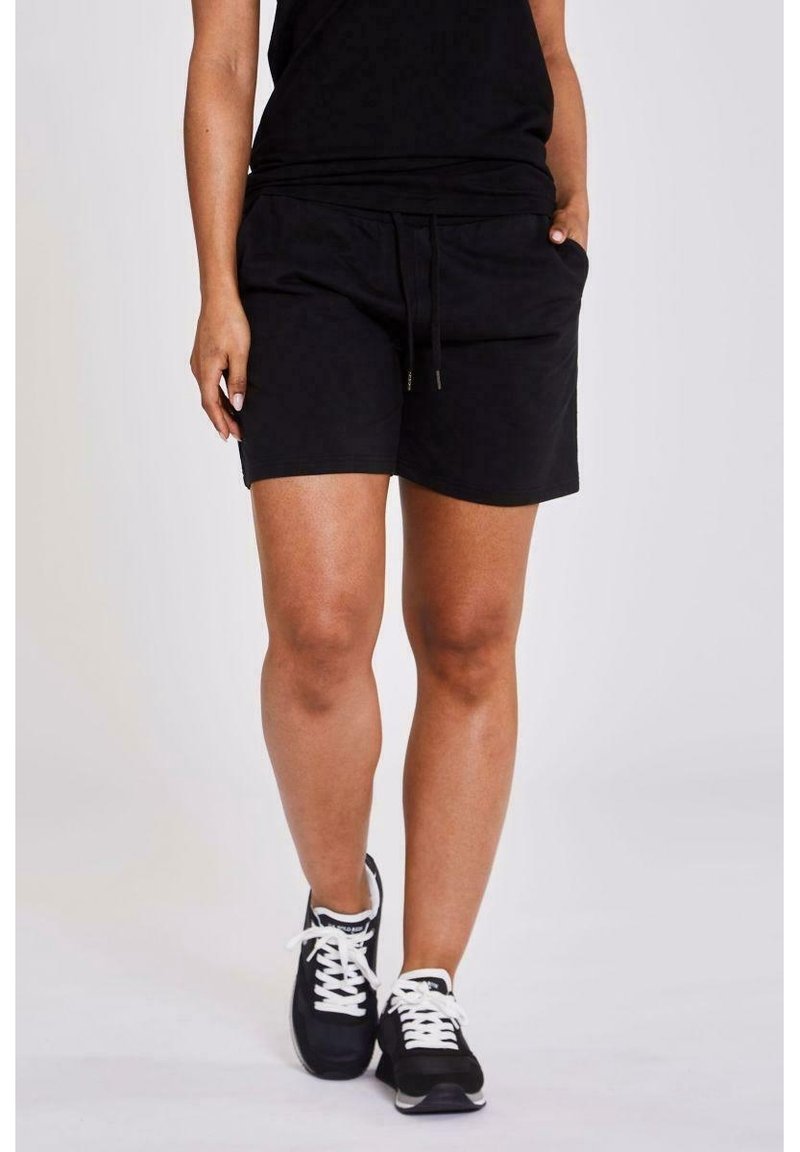 Personne portant un short noir à cordon, un haut noir et des baskets noires à lacets blancs, debout devant un fond uni.