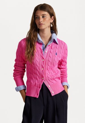 Cardigã de malha rosa com decote em V, com botões na frente e punhos canelados. Usado sobre uma camisa azul clara com gola.