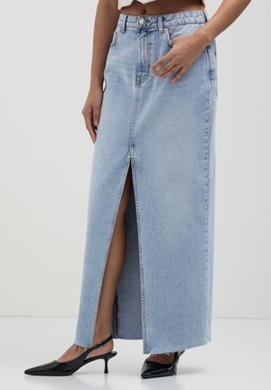 Frau trägt einen hellblauen Jeans-Maxirock mit Frontschlitz, kombiniert mit schwarzen, spitz zulaufenden Slingback-Absätzen, steht vor einem schlichten Hintergrund.