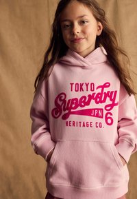 Ružová mikina s veľkým červeným potlačou s nápisom "TOKYO Superdry JPN 6 HERITAGE CO." Obsahuje predné vrecko a ribbované manžety.