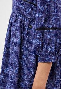 Μωβ φόρεμα με μοτίβα, που διαθέτει μπλε σχέδια παϊέτας, υφασμάτινη υφή, φουσκωτά μανίκια και ένα κουμπί στη στενή περιοχή του σώματος.