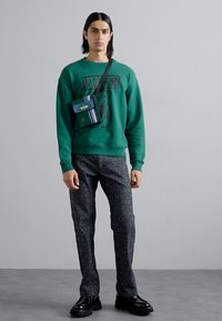 Sweatshirt verde cu design de urs, asortat cu pantaloni negri cu un model care afișează numele brandului. Accesoriu: geantă mică verde cu dungi.