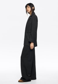 Traje de cuadros oscuros que incluye una americana larga y pantalones de pierna ancha. El material parece texturizado con un sutil patrón de cuadros. Destaca por su solapa.
