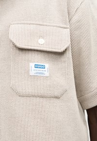 Μπεζ υφασμάτινο πουκάμισο με κοντά μανίκια, μπροστινή τσέπη με κουμπιά και λευκή ετικέτα με μπλε γράμματα «HUGO».
