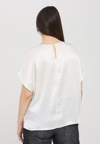 Blusa bianca di satin con maniche corte, apertura a chiave sul retro e una superficie liscia, caratterizzata da un orlo dritto e una vestibilità rilassata.