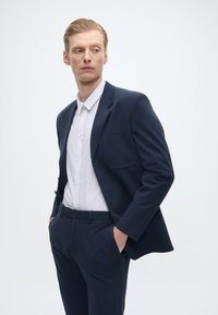 Tengerészkék öltönyzakó, kihajtott reverrel, egyetlen mellzsebbel, fehér gombos inggel és azonos tengerészkék nadrággal párosítva, minimalista design.