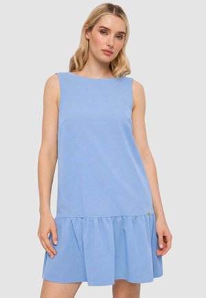 Donna con capelli biondi che indossa un vestito azzurro senza maniche con orlo arricciato, in piedi davanti a uno sfondo grigio uniforme.